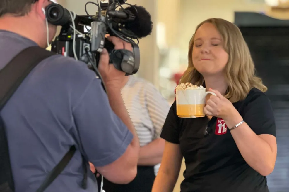 "Blonde woman smiling and sipping coffee while looking directly at the camera.